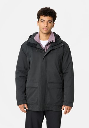 Young man wearing a black hooded winter jacket layered over a purple fleece jacket, standing against a plain light background.