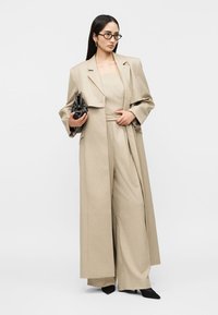 Woman with long dark hair wearing beige long coat, wide-leg pants, black pointed shoes, glasses, and holding a black handbag.