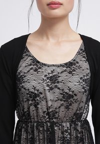 Mustap cardigan mustan ja valkoisen kukkalahkeisen pitsimekon päällä, jossa on istuva vyötärö, pyöreä pääntie ja läpinäkyvä kukkakuosi.