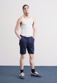 Débardeur blanc à côtes, shorts navy, baskets navy avec accents blancs. Le modèle se tient de manière décontractée, les mains dans les poches, devant un fond uni.
