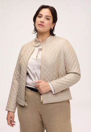 Kvinde iført en beige quiltet jakke over en lyse satinbluse og beige bukser, stående mod en ensfarvet lys baggrund.