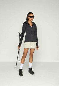 Črna zip-up jakna, svetli kratki hlače, črne pohodniške čevlje in trekking palica. Dodatki vključujejo temne sončne očala in bele nogavice.