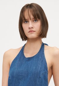 Denim halterneck topp med blå vaske, ermeløs design og buet kant. Stoffet har en glatt tekstur med synlige sømmer.