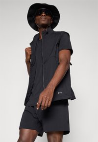 Black lightweight vest with zip closure, paired with matching t-shirt and shorts. Model wearing a bucket hat and sunglasses.