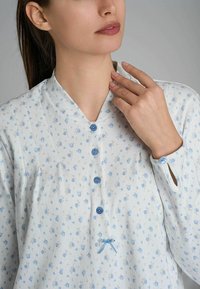 Top bianco con piccolo motivo floreale blu, chiusura frontale con bottoni, colletto alla coreana e fiocco decorativo. Presenta bottoni blu e polsini con bottone.
