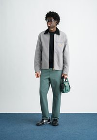 Grijze jas met zwarte kraag, zwart shirt, groene broek en zwarte schoenen. Draagt een kleine groene tas. Zonnebril weerkaatst licht.