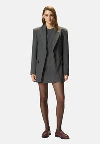 Blazer gris con solapas afiladas, cierre de un solo botón y bolsillos delanteros, combinado con un vestido gris y zapatos granate. Tejido liso, diseño estructurado.
