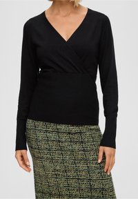 Czarna bluzka z długim rękawem i dekoltem w serek, o kroju kopertowym. Materiał wydaje się gładki i lekko dopasowany, zestawiony z wzorzystą zieloną spódnicą.
