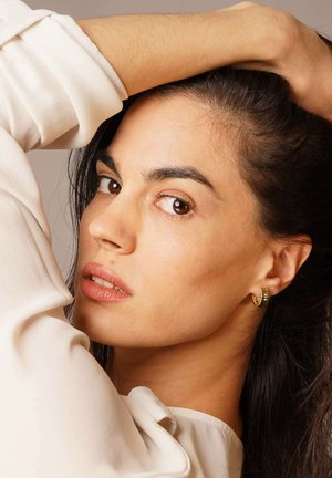 Model with long dark hair, wearing a gold hoop earring. She has a pale skin tone and is draped in a light fabric blouse.