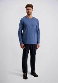 Ein Mann steht in einem blauen Langarmhemd, schwarzen Hosen und dunkelbraunen Slippern vor einem schlichten, hellen Hintergrund.