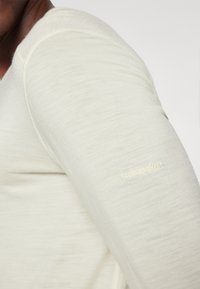 Chemise à manches longues beige clair en tissu doux, arborant un design ajusté et un petit logo brodé en or sur la manche.
