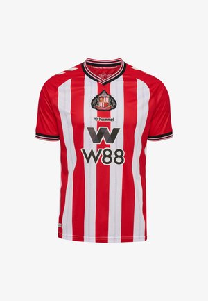 Piłkarska koszulka w czerwono-białe paski wykonana z lekkiego materiału, z czarnym kołnierzykiem, logo drużyny oraz wyraźnym czarnym napisem sponsora.