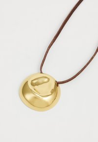 Ciondolo d'oro con superficie liscia e arrotondata, appeso a una corda in pelle marrone. Il design è minimalista e presenta una finitura lucida.