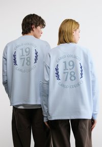 Δύο άτομα φορούν ανοιχτόχρωμες μπλούζες με κουκούλα με το κείμενο "CARLO COLUCCI 1978" και σχέδια στεφάνου δάφνης στην πλάτη, στέκονται δίπλα-δίπλα.