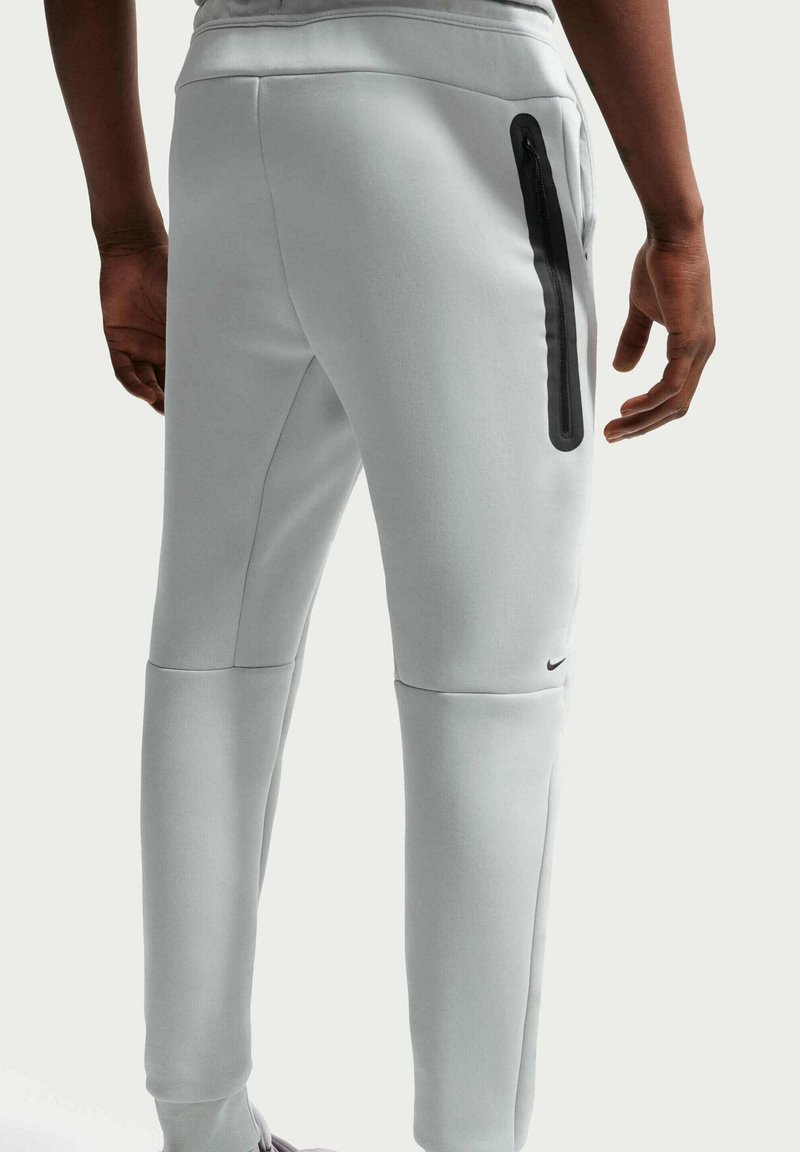 Person trägt hellgraue Sporthose mit einer schwarzen Reißverschlusstasche und kleinem schwarzem Logo nahe dem Knie, steht mit entspannten Händen.