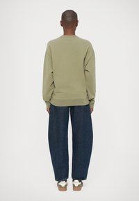 Sudadera verde oliva con hombros caídos, puños y dobladillo acanalados, combinada con unos vaqueros de pierna ancha azul oscuro y zapatillas blancas. Vista trasera.