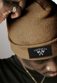 Brown ribbed knit beanie featuring a black label with "valhalla" and ax graphic, close-up on a person's head with hand partially visible.