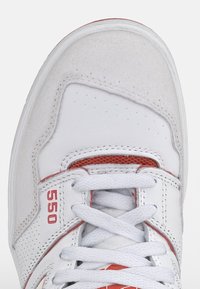 Baskets blanches en matériaux suédé et cuir, avec des accents rouges sur les côtés et la languette, des détails perforés et des lacets plats blancs. Numéro de modèle 550.