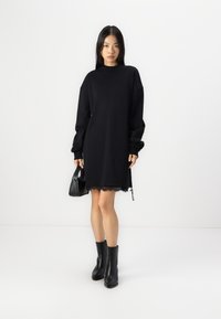 Rochie supradimensionată neagră din molton, cu dantelă la margine, mâneci lungi și guler înalt. Asortată cu cizme negre până la gleznă și o poșetă mică.