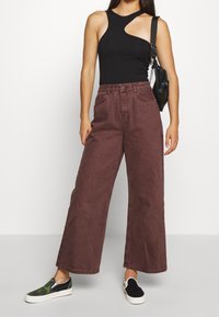 Mujer con una blusa sin mangas negra asimétrica, pantalones marrones de pierna ancha, zapatillas de andar por casa desiguales y un bolso negro de hombro.