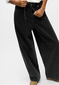 Pantalones de mezclilla negros de pierna ancha y cintura alta, con un lazo ajustable y detalles de costura en contraste. Textura suave.
