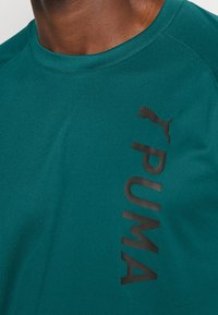 Tyrkysové sportovní tričko s krátkými rukávy, s černým logem "PUMA" a siluetou na přední straně, vyrobené z hladkého, lehkého materiálu.