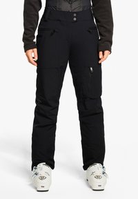 Schwarze Skihosen mit strukturierter Oberfläche, ausgestattet mit mehreren Taschen, Reißverschluss und schmaler Passform. Tragen mit weißen Skischuhen.