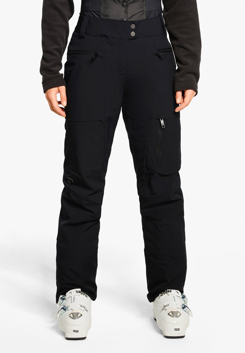 Schwarze Skihosen mit strukturierter Oberfläche, ausgestattet mit mehreren Taschen, Reißverschluss und schmaler Passform. Tragen mit weißen Skischuhen.