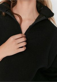 Sudadera negra con cremallera, que presenta un cuello alto, una textura suave y un diseño ajustado. Mano sosteniendo el tirador de la cremallera en la parte frontal.