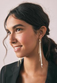 Dolgi srebrni uhani z teksturiranimi verigami in majhnimi srčastimi detajli, ki elegantno visijo. Nosijo jih model z temnimi lasmi in črnim blazerjem.