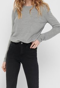 Hellgrauer Pullover mit Textur und langen Ärmeln, kombiniert mit hoch taillierten schwarzen Jeans mit Vordertaschen und Knopfverschluss.