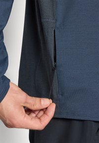 Navy long-sleeve shirt with a zipper side pocket, textured fabric, and thumbholes at the cuff. A hand is pulling at the zipper tag.
