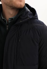 Giacca puffer blu navy con tasca a zip sul petto, collo alto e cappuccio attaccato, caratterizzata da un design trapuntato e una texture liscia.