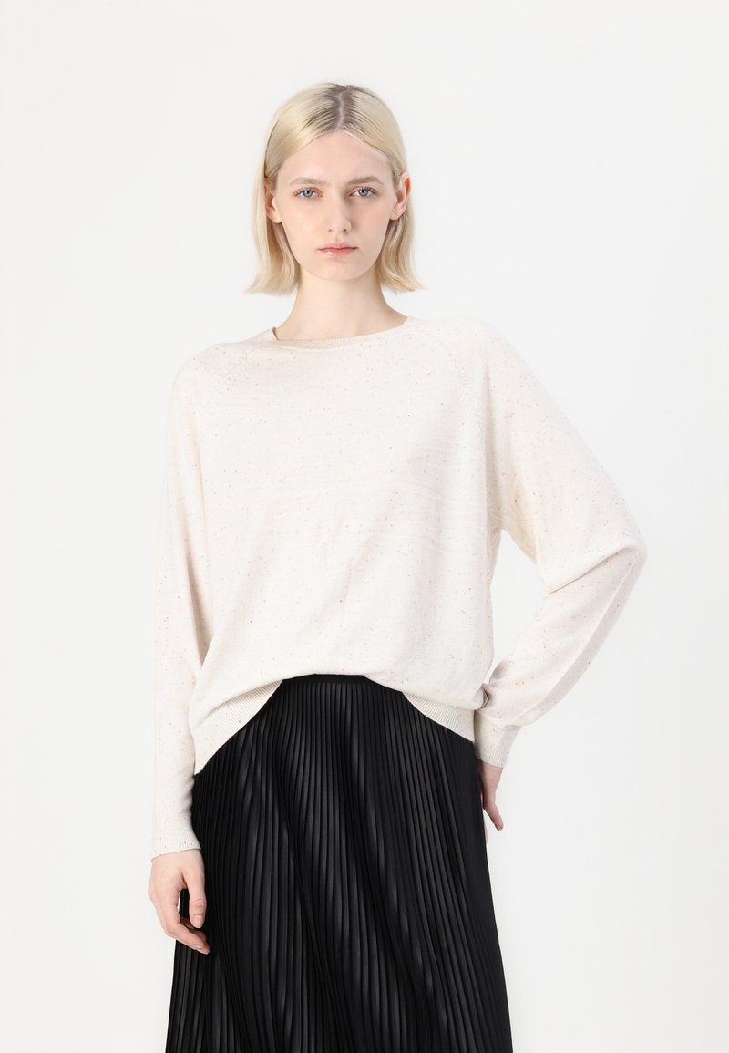 Hellbeige, langärmliges Strickoberteil mit gesprenkelter Textur, weitem Ausschnitt und leicht verlängerter Rückseite, kombiniert mit einem schwarzen, plissierten Rock.