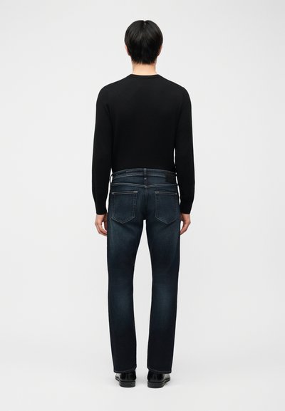 Άτομο που στέκεται με την πλάτη στραμμένη, φοράει μαύρο μπλουζάκι με μακριά μανίκια, σκουρόχρωμο μπλε τζιν με πίσω τσέπες και μαύρα παπούτσια σε απλό λευκό φόντο.