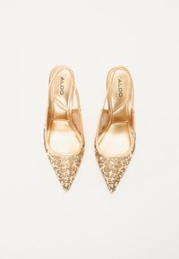 Gule, spisse slingback-hæler med utskåret blomsterdesign og en blank finish. Merkenavnet "ALDO" synlig på innersålen.