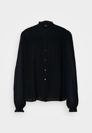 Svart blus med hög krage, långa genomskinliga ärmar, knappstängning framtill och texturerad tyg med prickar. Har samlad detalj längs oket.