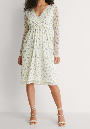 Woman wearing knee-length cream dress with blue floral print and sheer long sleeves, paired with beige strappy heels.