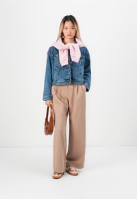 Young woman wearing a denim jacket, beige wide-leg pants, pink sweatshirt draped over shoulders, holding a brown handbag, standing on white background.