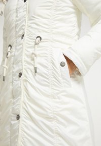 Doudoune blanche avec texture froncée, poches avant, cordons de serrage ajustables et accents métalliques, véhiculant un design épuré et fonctionnel.