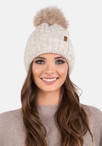 Bonnet en tricot crème avec texture côtelée, présentant un grand pompon en fausse fourrure sur le dessus et une petite étiquette en cuir.