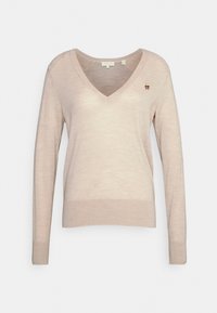 Pull long beige clair à manches longues avec un décolleté en V, fabriqué dans un tissu doux, comportant une petite épingle décorative en forme de fleur sur le côté gauche.
