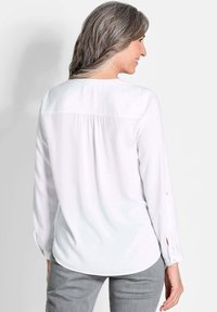 Weiße Bluse mit langen Ärmeln, runder Saumlinie, gestepptem Rückendetail und geknöpften Manschettenärmel. Getragen mit grauen schmalen Hosen.