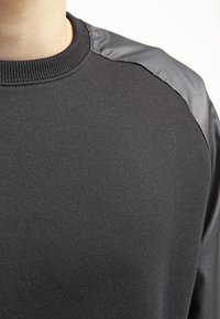 Sort sweatshirt med en tekstureret strikket krop og skinnende sort læderlignende materiale på skuldrene. Har en ribbet halsudskæring.