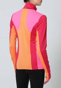 Koszulka sportowa z długim rękawem wykonana z mieszanki jasnoróżowych, pomarańczowych i koralowych paneli, z wysokim kołnierzem i gładkim, elastycznym materiałem.