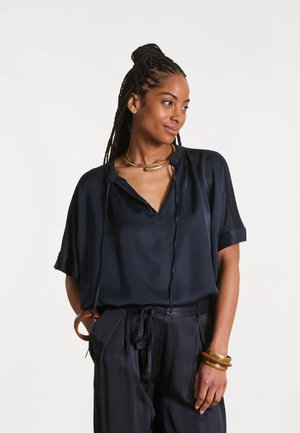 Femme aux cheveux tressés portant un chemisier et un pantalon en satin noir, accessoirisée d'un collier et de bracelets en or, regardant sur le côté les mains dans les poches.