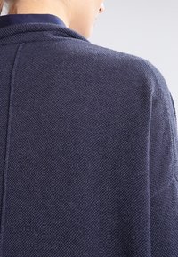 Marinblå stickad cardigan med texturerad yta, med design för nedvikt axel och ribbad halsringning. Subtila sömnadsdetaljer är synliga.