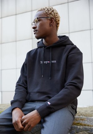 Sweatshirt à capuche noir avec le logo "Tommy Jeans" en blanc et rouge, associé à un pantalon gris. Tissu lisse, cordons de serrage et poignets côtelés.