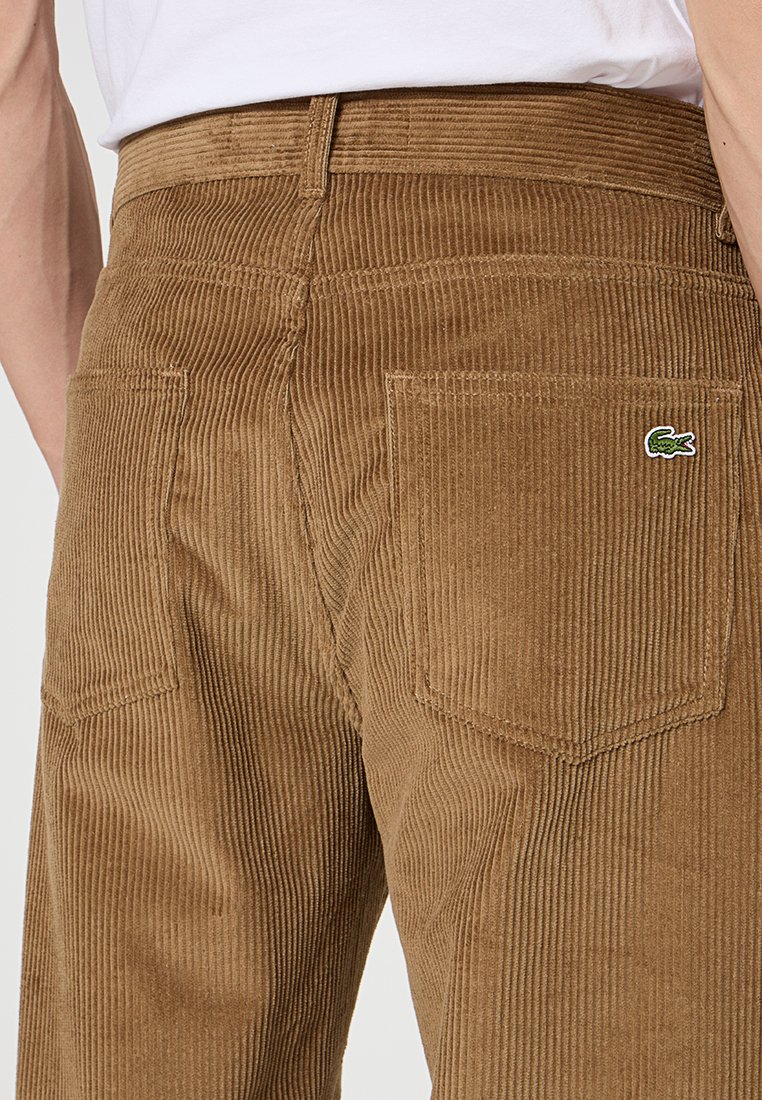 Pantalon en velours côtelé marron avec des rayures verticales, doté de deux poches arrière et d'un petit logo vert Lacoste sur la poche droite.