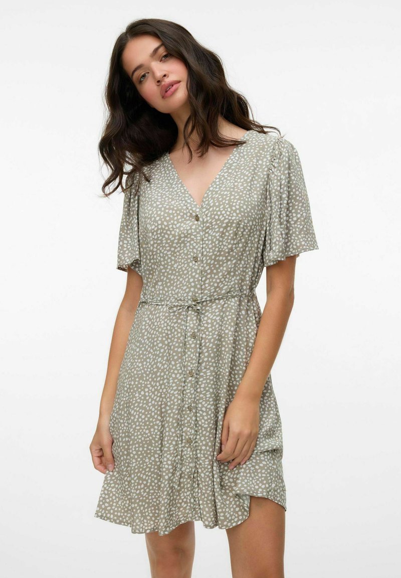 Robe verte à manches courtes avec un décolleté en V, boutons sur le devant, et pois blancs. Tissu léger avec une taille cintrée et une jupe évasée.